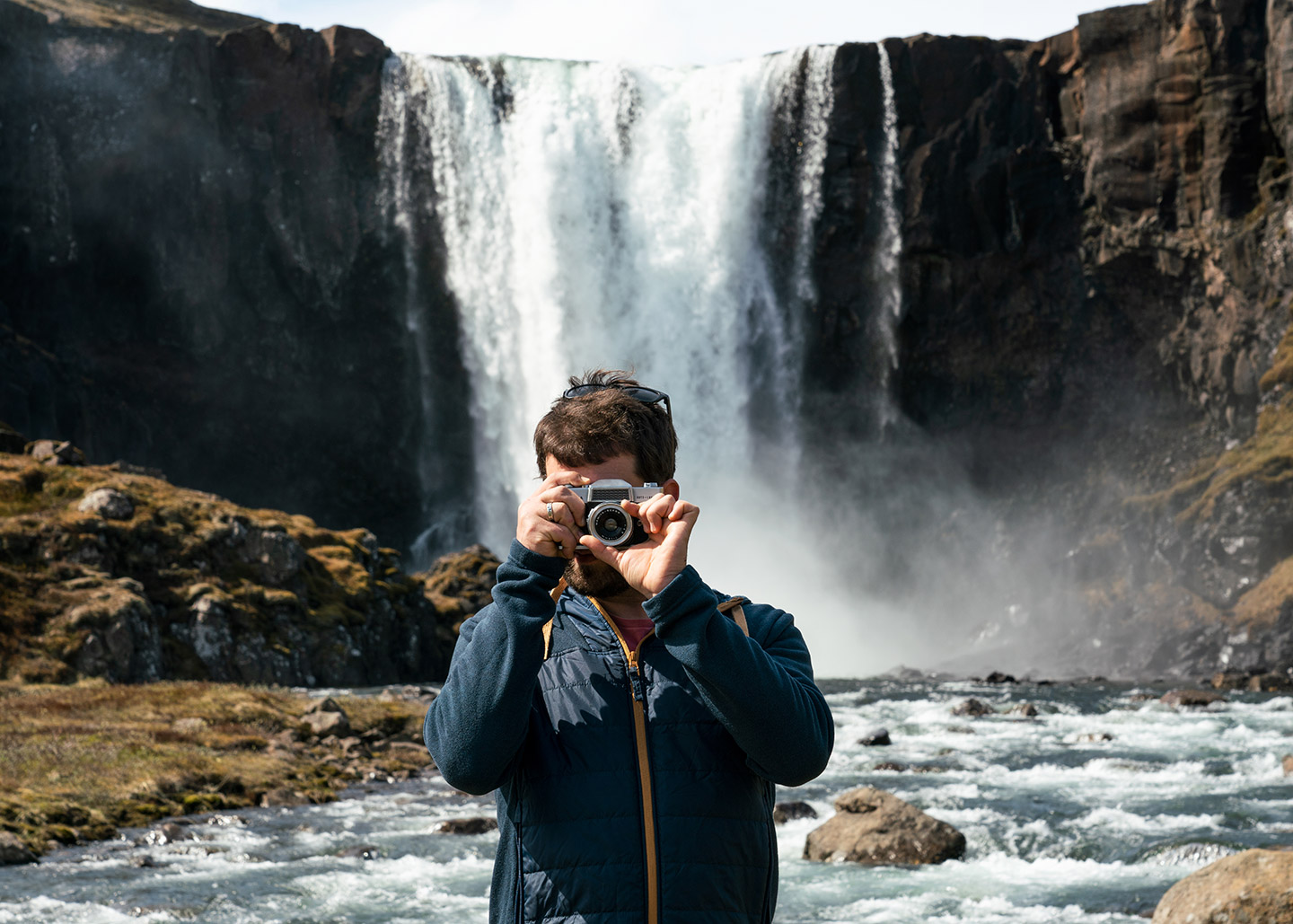 A man is standing in front of Gufufoss waterfall in East Iceland, holding a camera and taking a photo