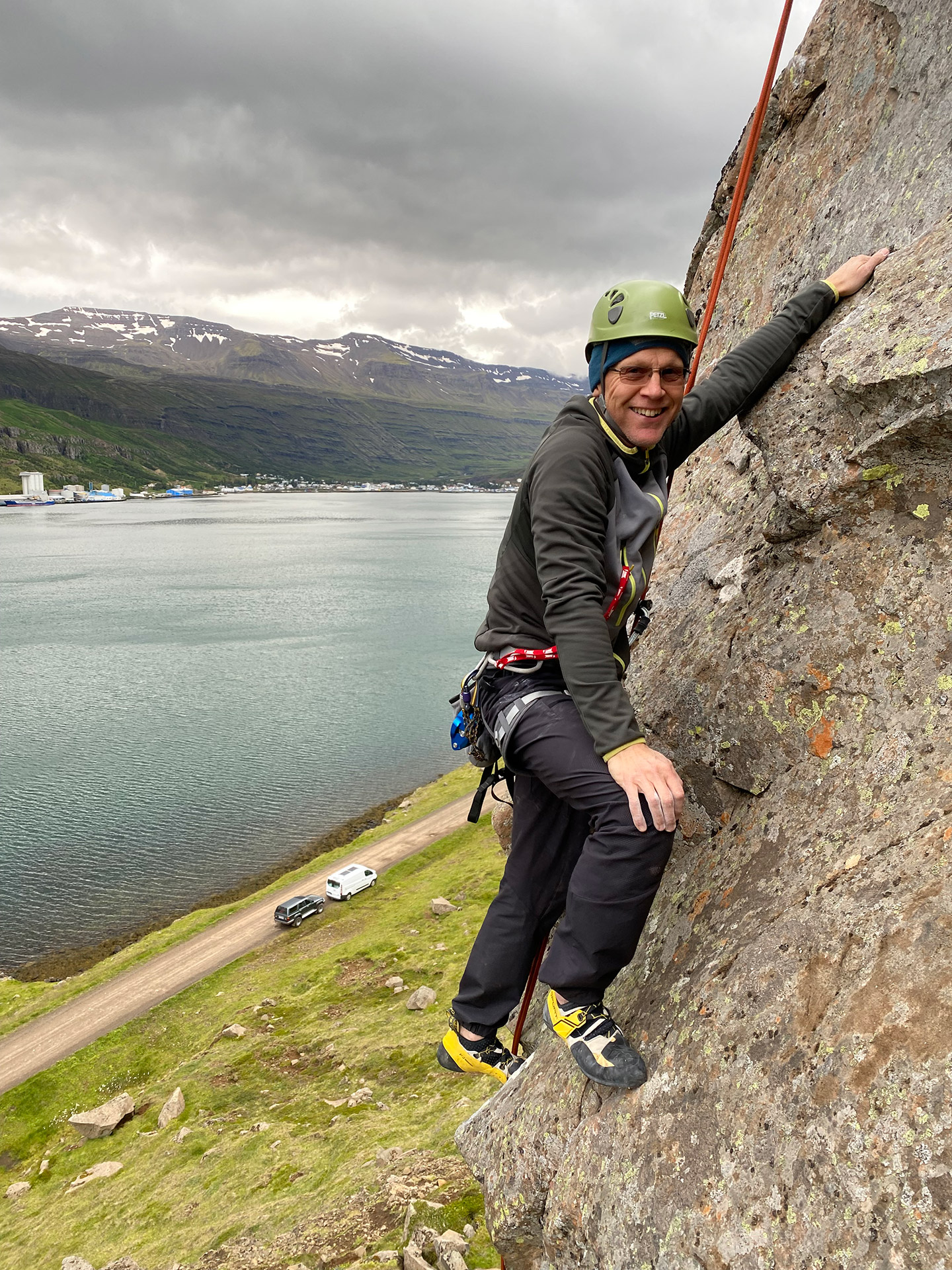 A happy climber working his way up the cliff at Arnarklettar in Seyðisfjörður with an overview of the town in the background.