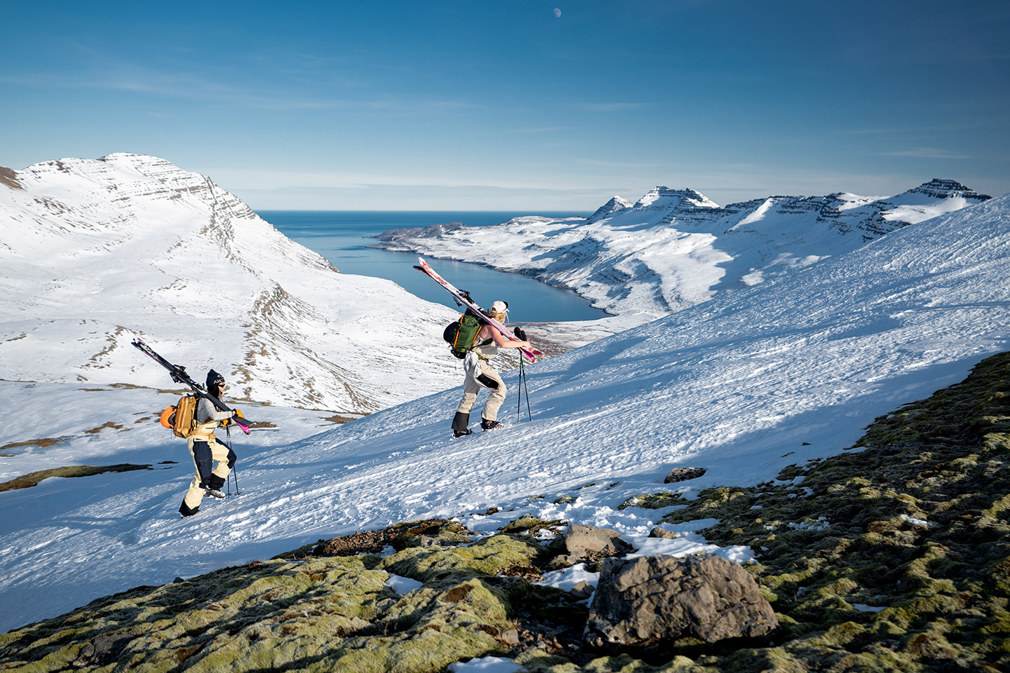 Two skiers are hiking up a snowy mountain in East Iceland. In the background, a beautiful fjord can be seen, with its deep blue waters extending out to the horizon.