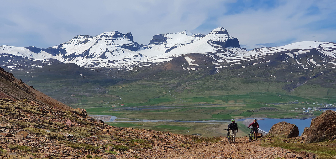 Mt bikers hiking up a trail with their bikes. In the background there is a magnificent view of Dyrfjöll